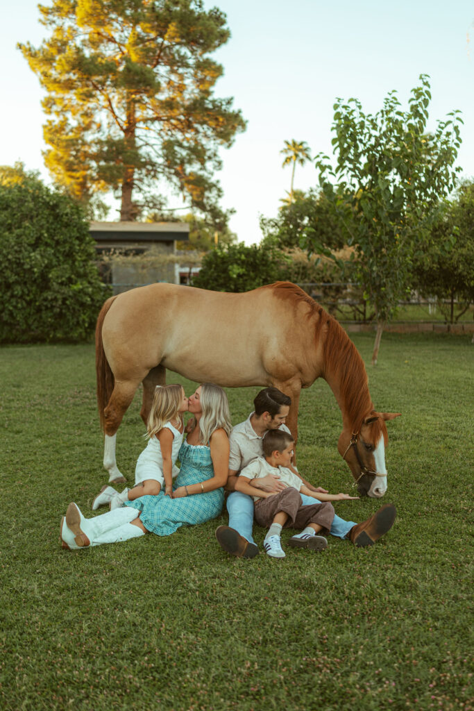 Family maternity photographer in Gilbert capturing an expecting family relaxing beneath their horse during a golden hour pregnancy announcement session at Floyd Family Farms.