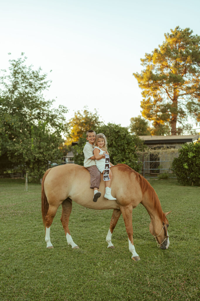 family photography featuring siblings sitting together on their family horse while holding ultrasound photos during a pregnancy announcement session at Floyd Family Farms in Gilbert.