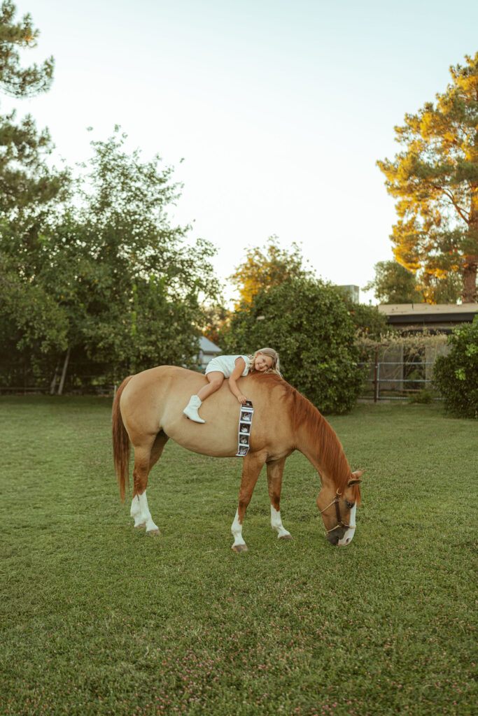 Gilbert maternity photographer capturing a little girl cuddling her family horse while ultrasound photos rest on the horse's back during a pregnancy announcement session at Floyd Family Farms.