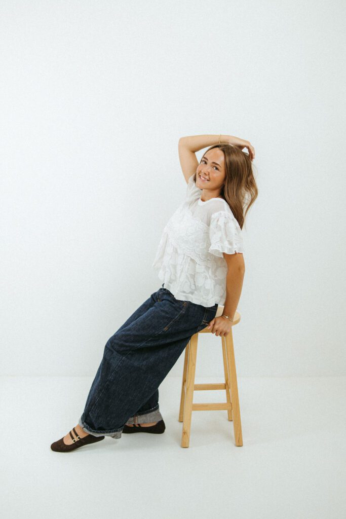 Young woman posing on a wooden stool in a white studio for her 18th birthday portraits in Queen Creek, Arizona.