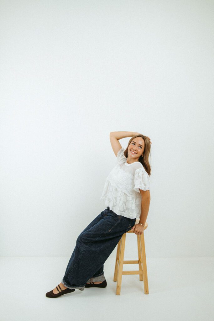 Young woman posing on a wooden stool in a white photography studio for creative 18th birthday portraits.