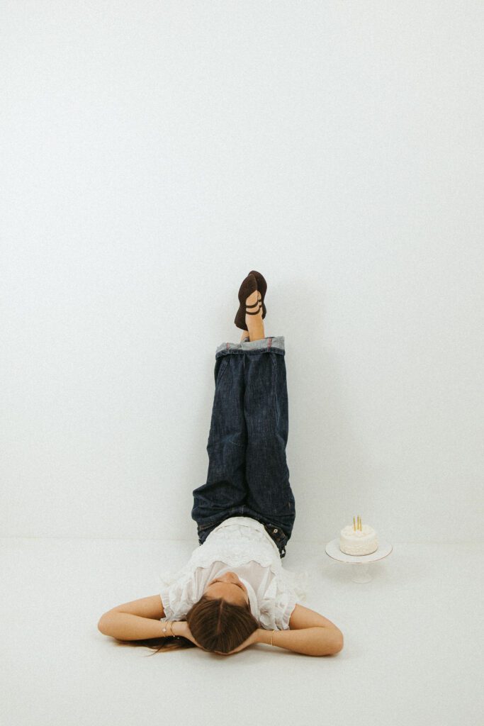Creative full-body portrait of a young woman lying on the studio floor with her legs raised beside a birthday cake.