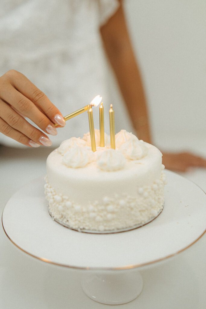 Close-up of an elegant white birthday cake with lit candles and a young woman's hand during an 18th birthday photo session.