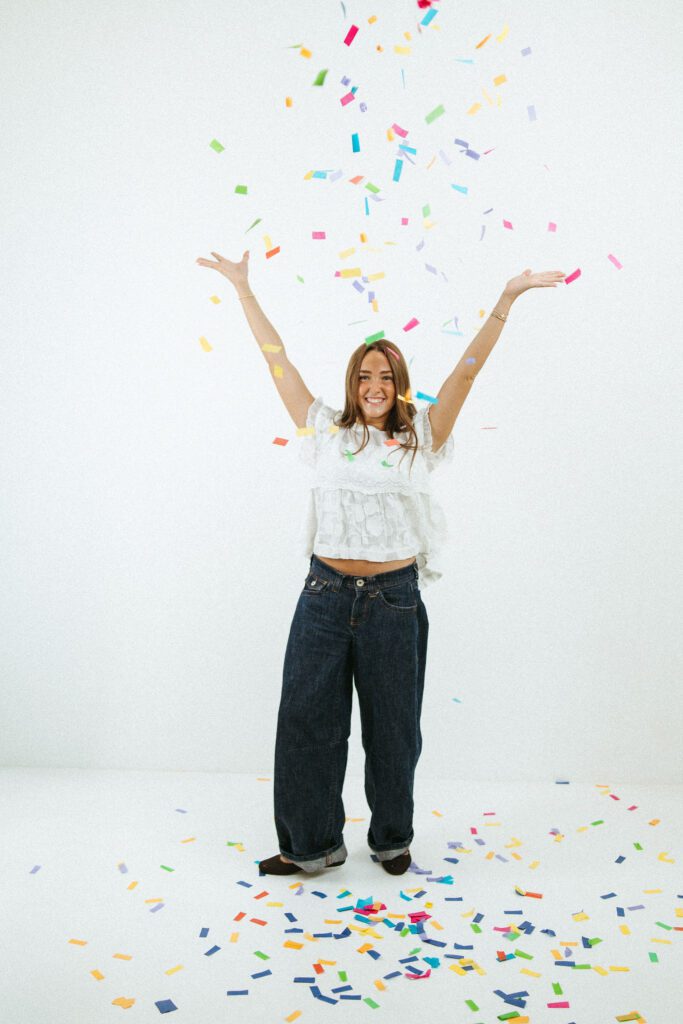 Full-body 18th birthday portrait of a young woman celebrating with colorful confetti in a white studio.