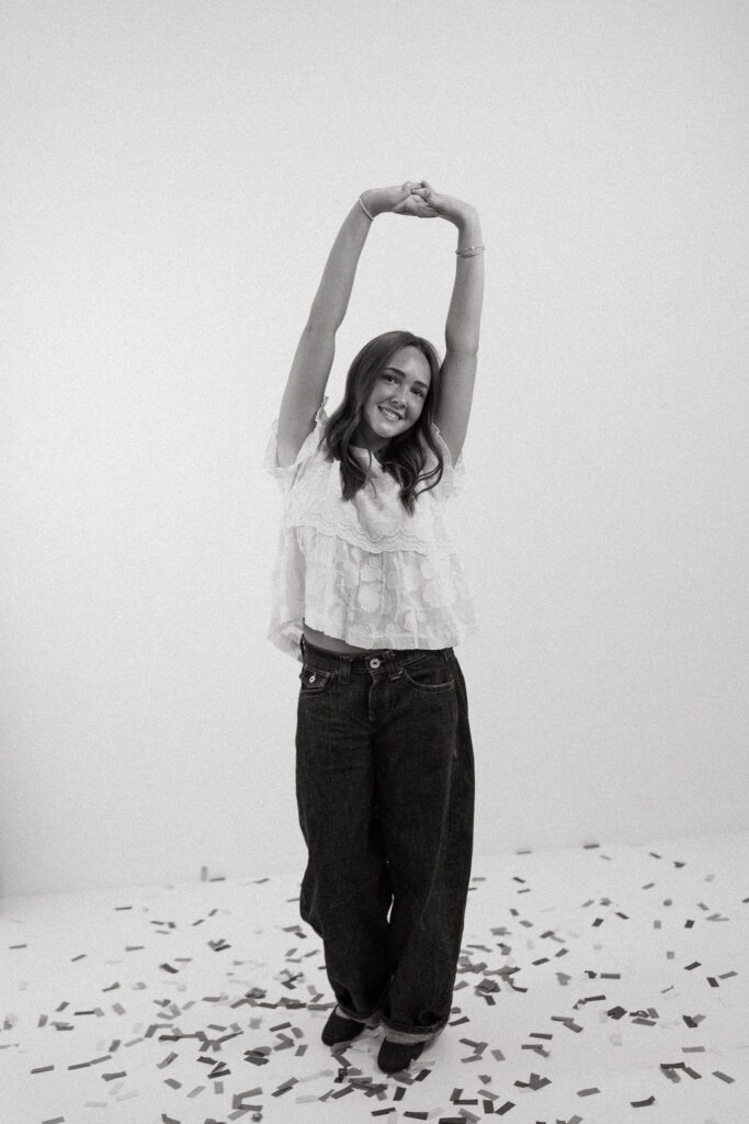 Black and white 18th birthday portrait of a young woman standing among confetti in a studio.