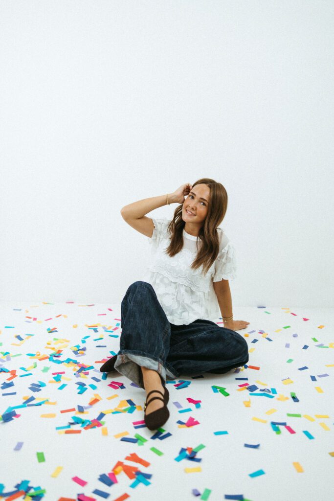 Young woman sitting casually among colorful confetti during her 18th birthday portraits in a Queen Creek photography studio.