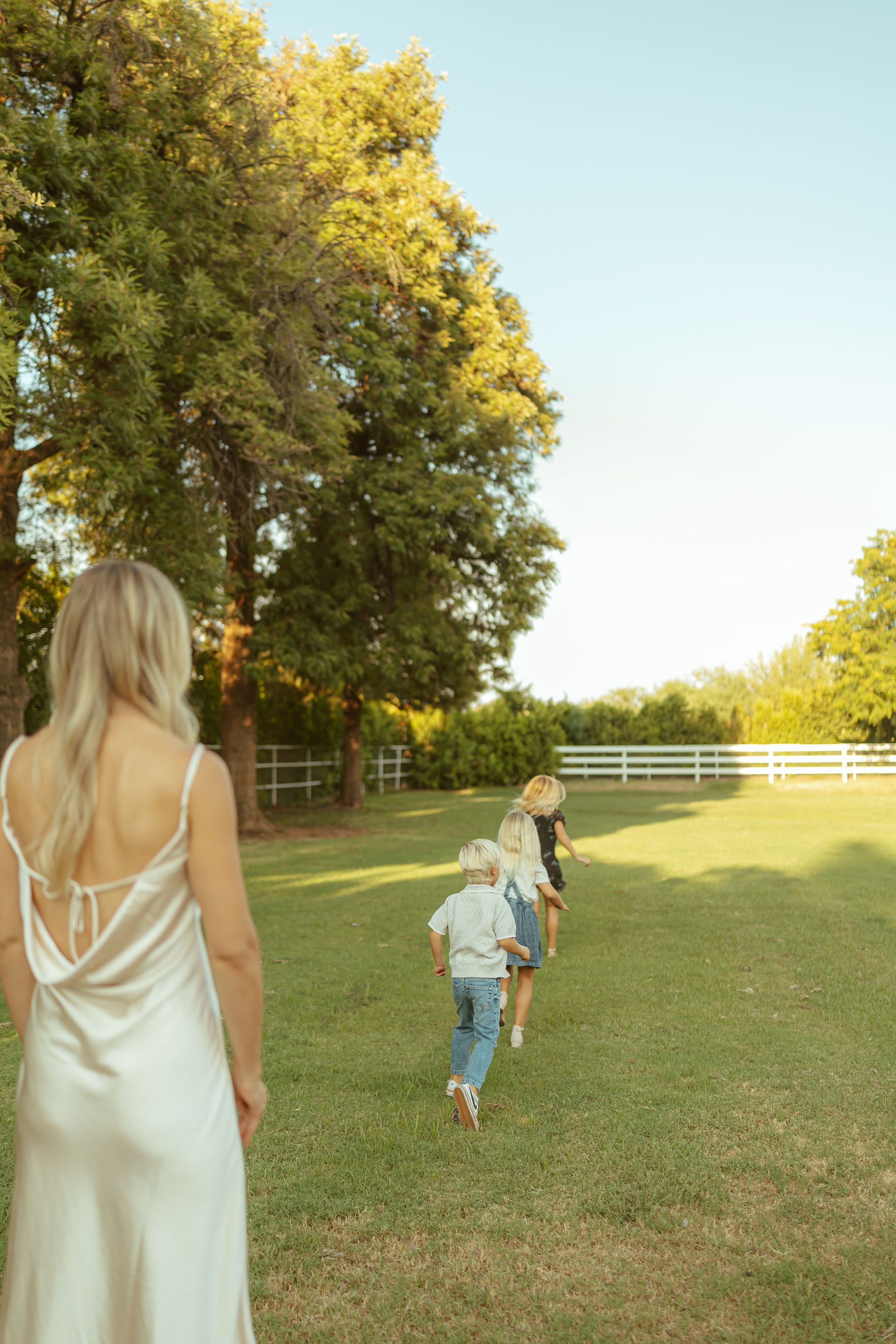 Queen Creek family photography featuring a mother in a silk slip dress watching her children run across an open lawn at a luxury farm venue.