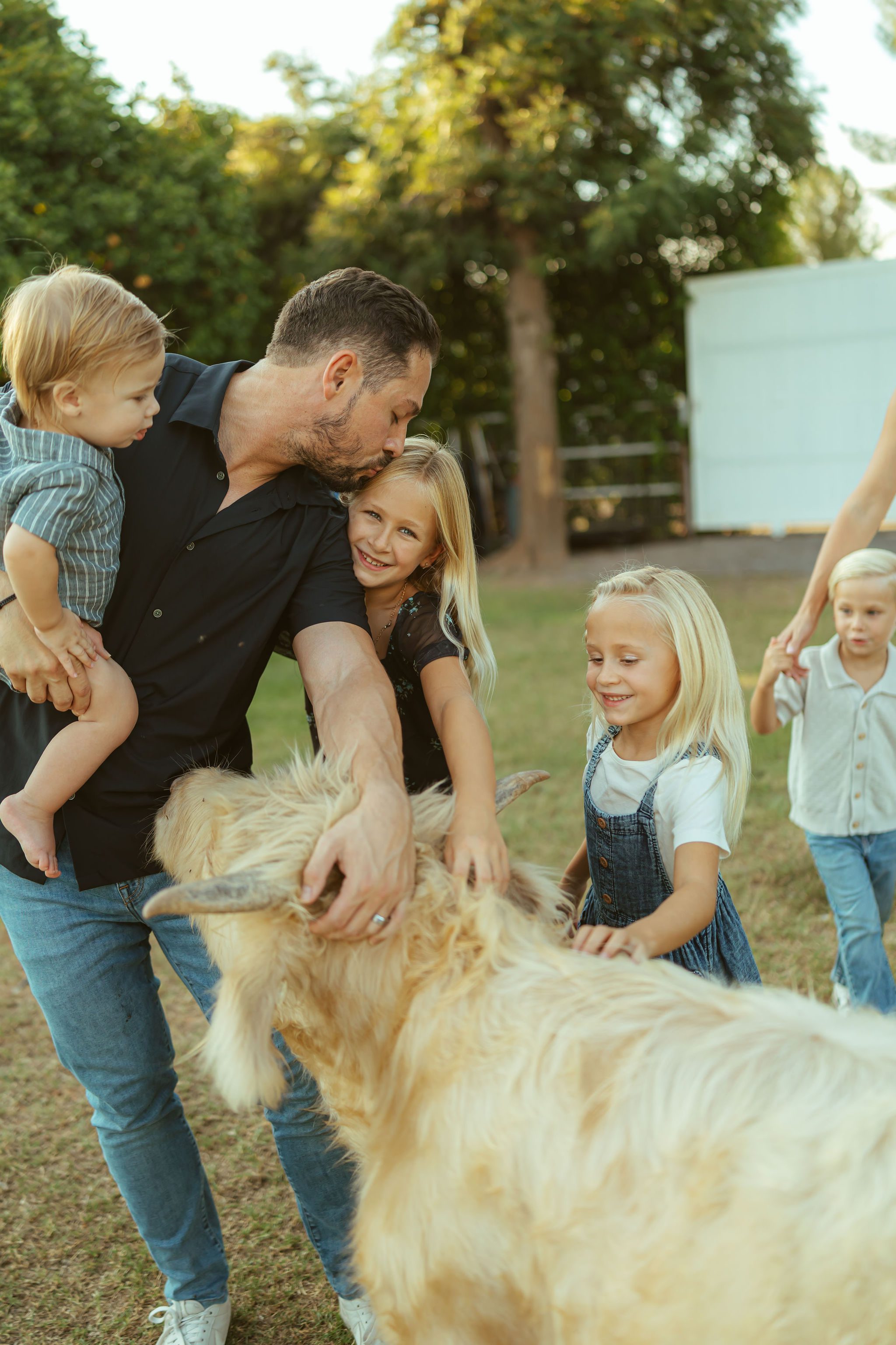 Mesa family photographer documenting a father kissing his toddler while his daughters pet a fluffy farm animal at an upscale country estate session.