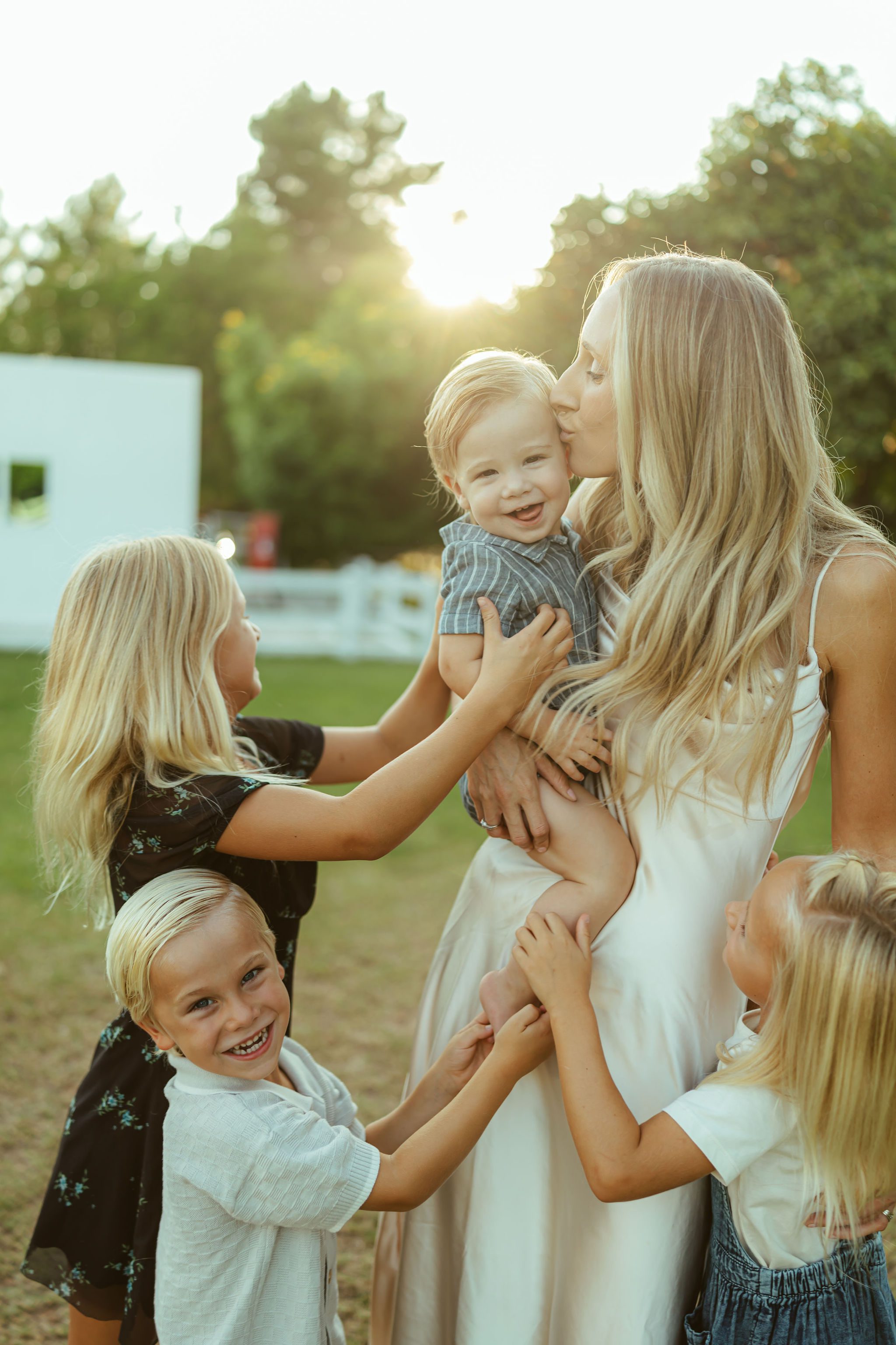 Phoenix family photographer capturing a sweet candid moment of a mother kissing her toddler boy while surrounded by her children at sunset.