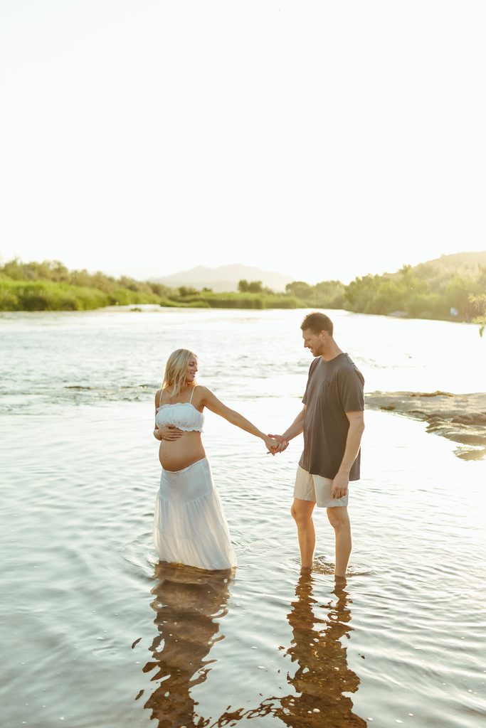 Pregnant woman in a white maternity dress standing in the Salt River with her partner