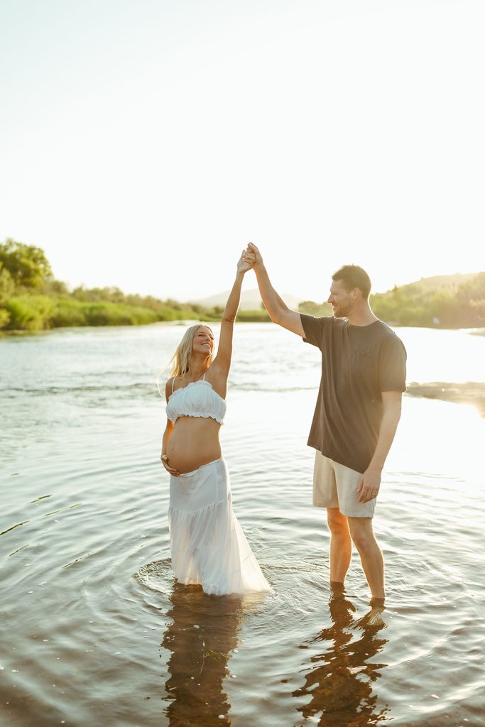 Expecting couple holding hands and raising their arms together in the Salt River at sunset