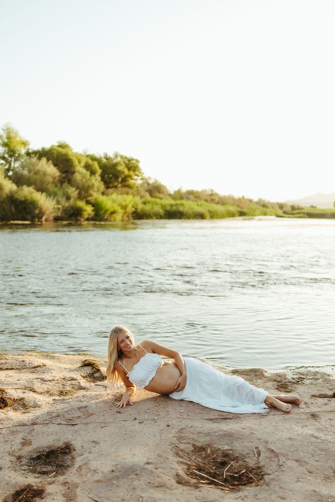 Pregnant woman in a white maternity dress reclining on the sandy shoreline of the Salt River in Mesa