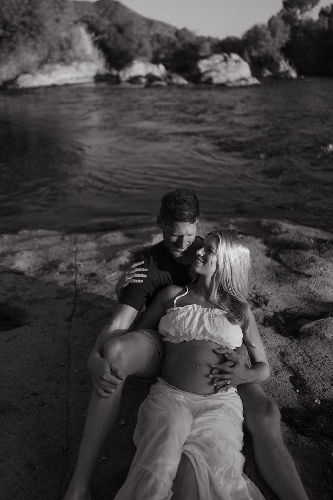 Black and white maternity portrait of an expecting couple sitting together beside the Salt River