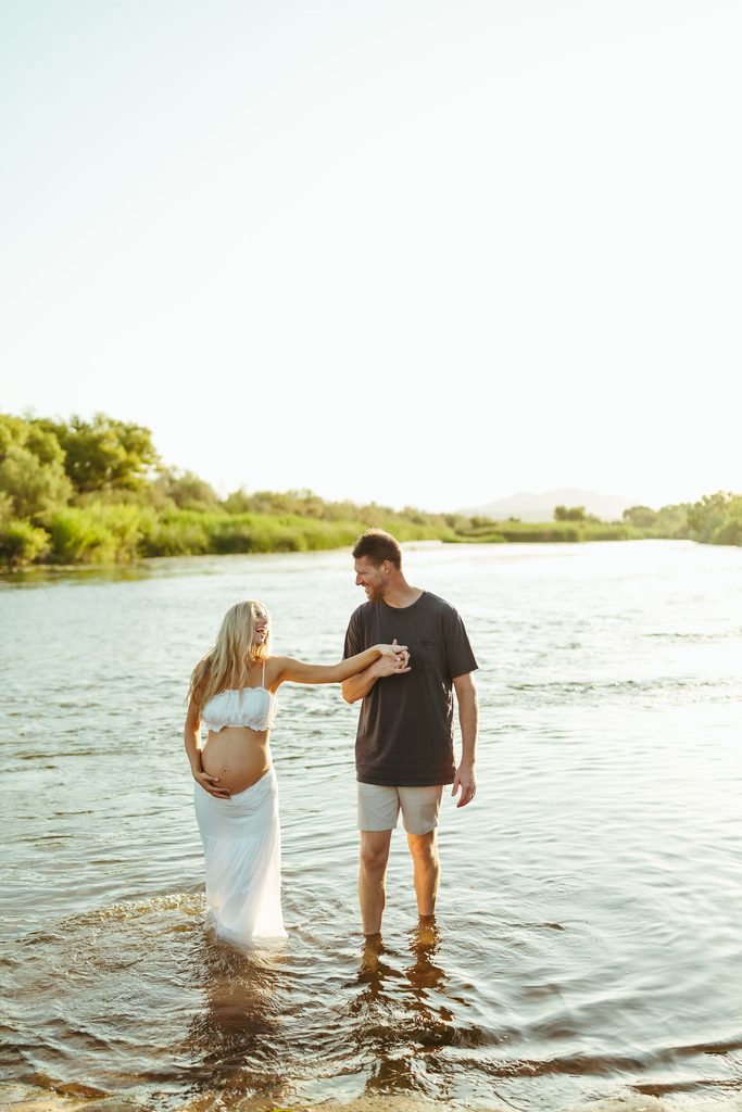 Expecting couple holding hands while walking through the Salt River during their Mesa maternity session