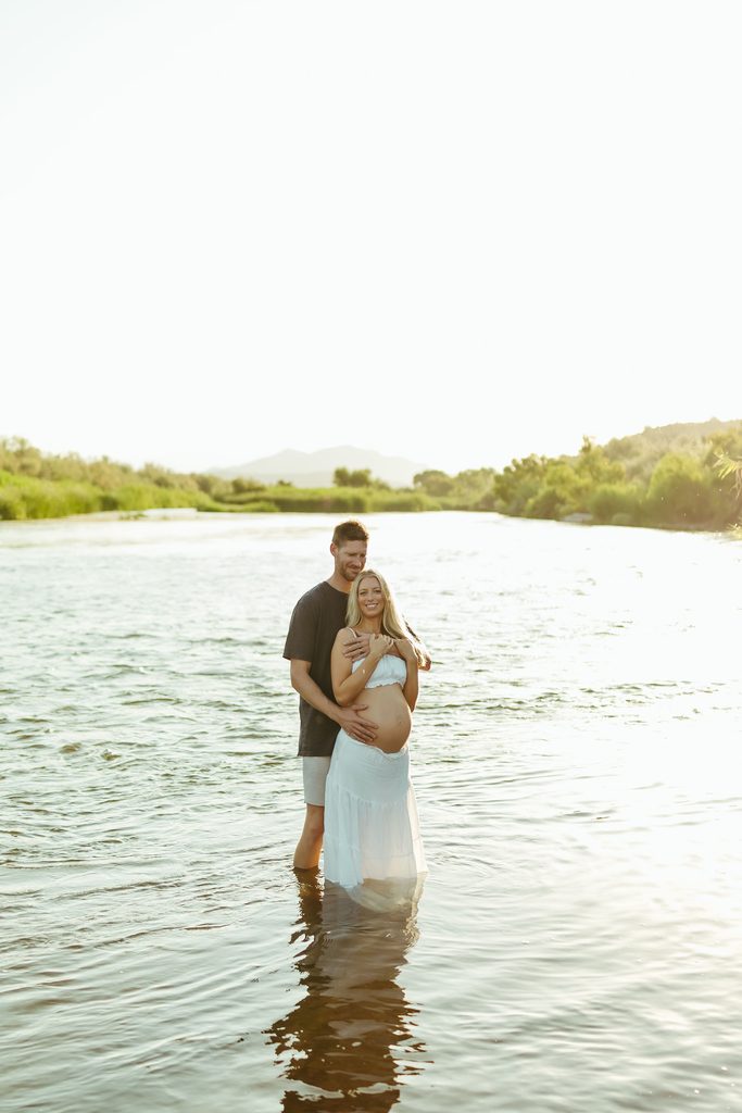 Pregnant woman in a white maternity dress standing in the Salt River with her partner