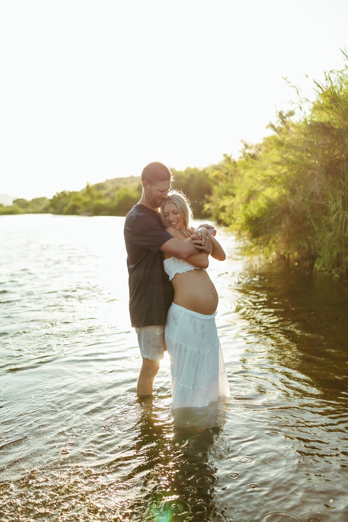 Pregnant couple standing together in the Salt River surrounded by greenery during golden hour