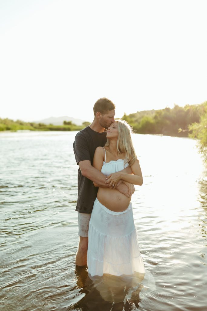Pregnant woman and her partner embracing in the Salt River at sunset during a Mesa, Arizona maternity session