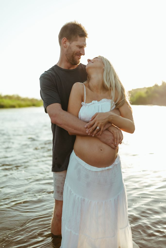 Black and white maternity portrait of a pregnant couple by the river, with the father kneeling beside the mother-to-be