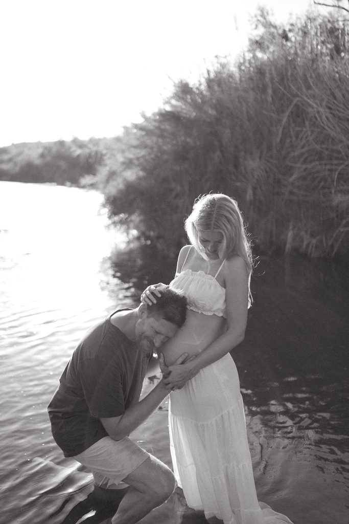 Pregnant couple embracing in shallow river water during a golden-hour maternity photography session
