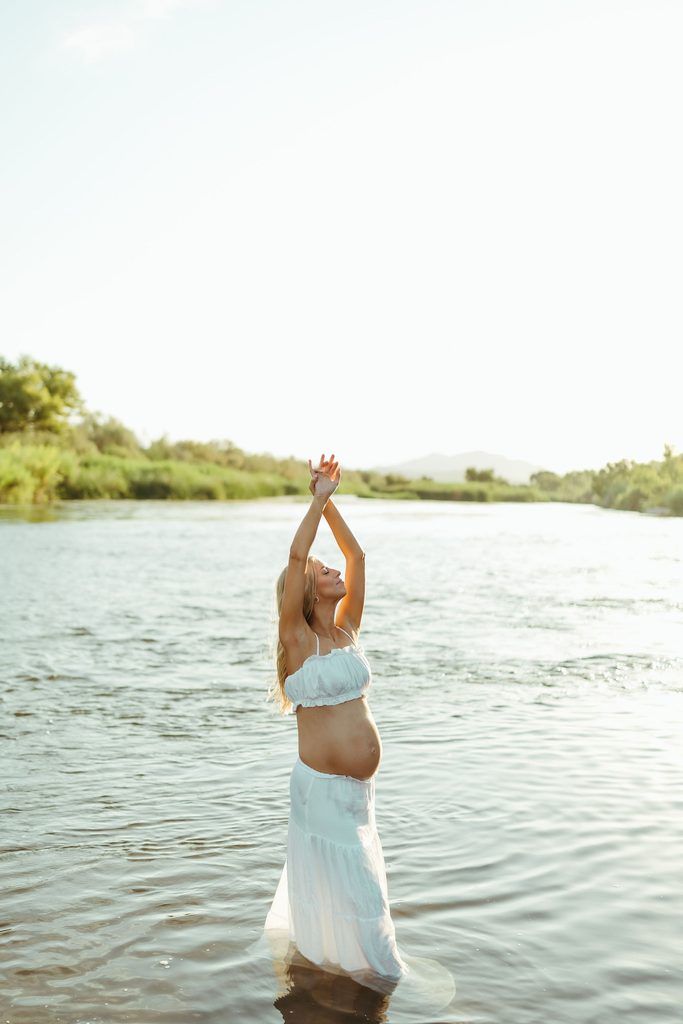 Pregnant woman standing in a river with her arms raised during a sunset maternity photo session