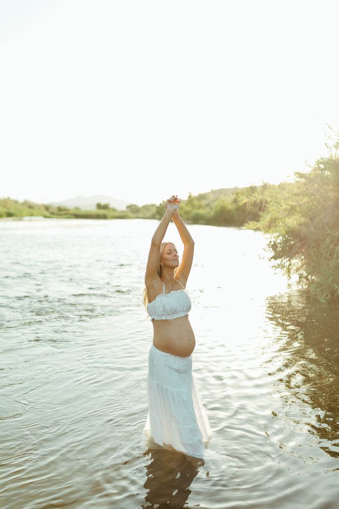 Pregnant woman in a white dress standing in the water with her arms raised during an Arizona maternity session