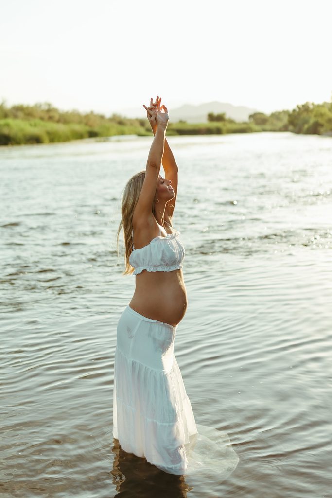 Pregnant woman in a white two-piece dress standing in a river with her arms raised during a golden-hour maternity session in Arizona