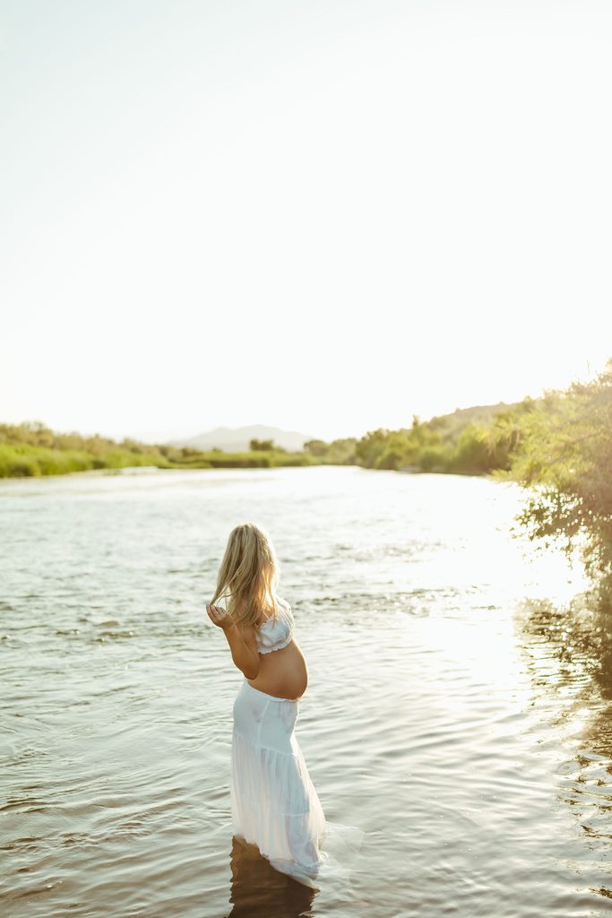 Pregnant woman standing in a river with her arms raised while wearing a flowing white maternity dress
