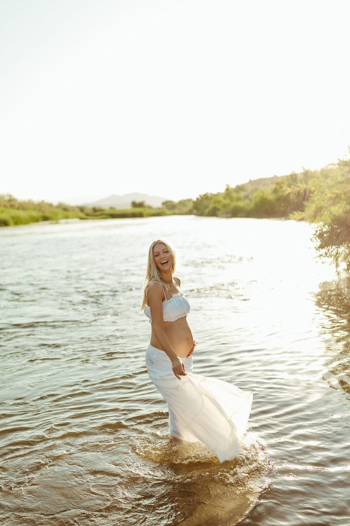 Pregnant woman in a flowing white dress walking through a river during a golden-hour maternity photography session in Arizona