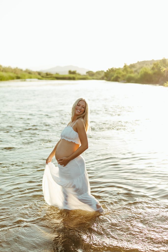 Pregnant woman in a white two-piece maternity outfit posing in the river at golden hour