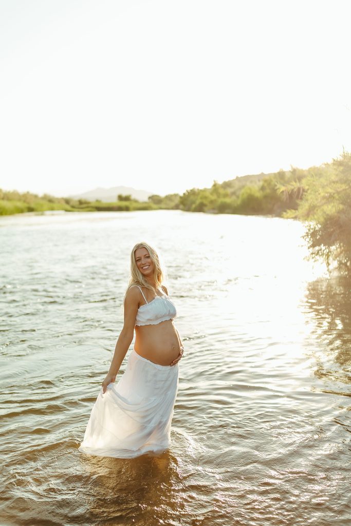 Pregnant woman smiling while standing in shallow river water during an outdoor Arizona maternity session