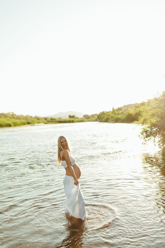 Pregnant woman in a white dress standing in a river surrounded by warm golden-hour light during her maternity photography session