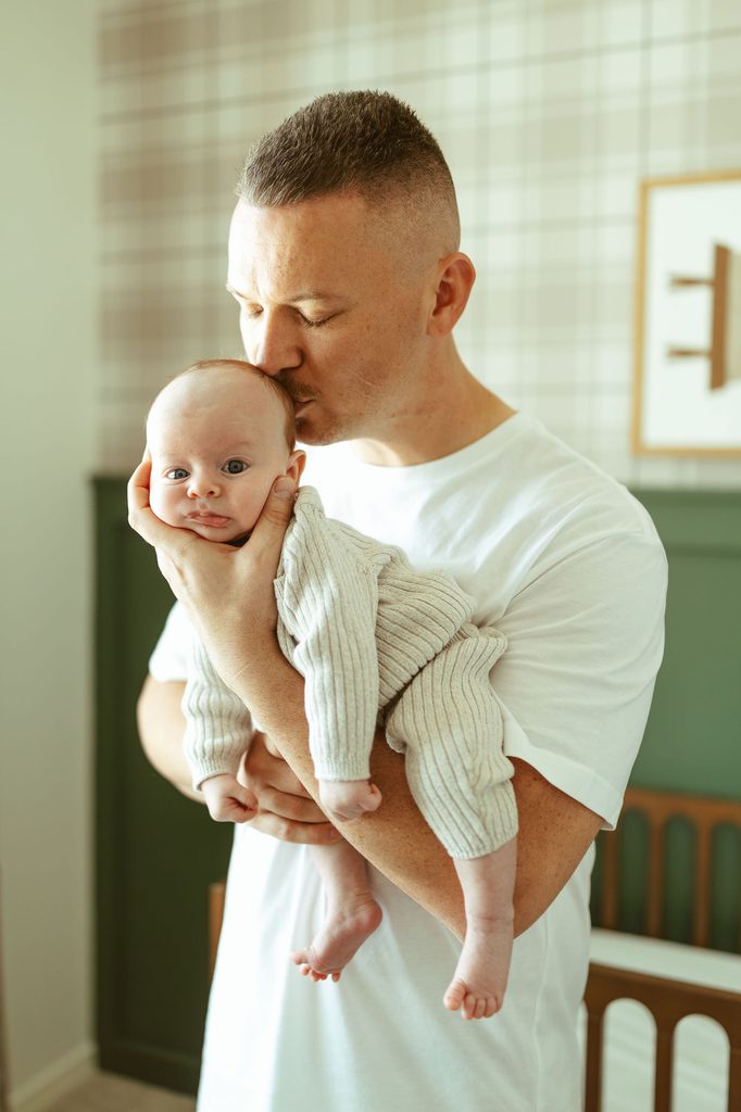 Gilbert newborn photographer capturing a father gently kissing his newborn baby while holding him in their Gilbert, Arizona home.