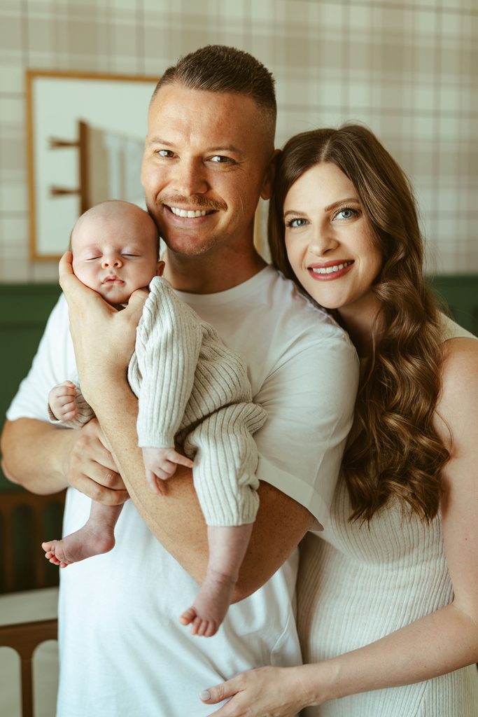 In-home newborn photography in Gilbert, Arizona featuring new parents smiling while holding their newborn baby together in the nursery.