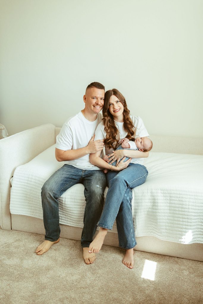 In-home newborn family photography in Gilbert, Arizona featuring new parents cuddling their baby together on a bright bedroom bed.
