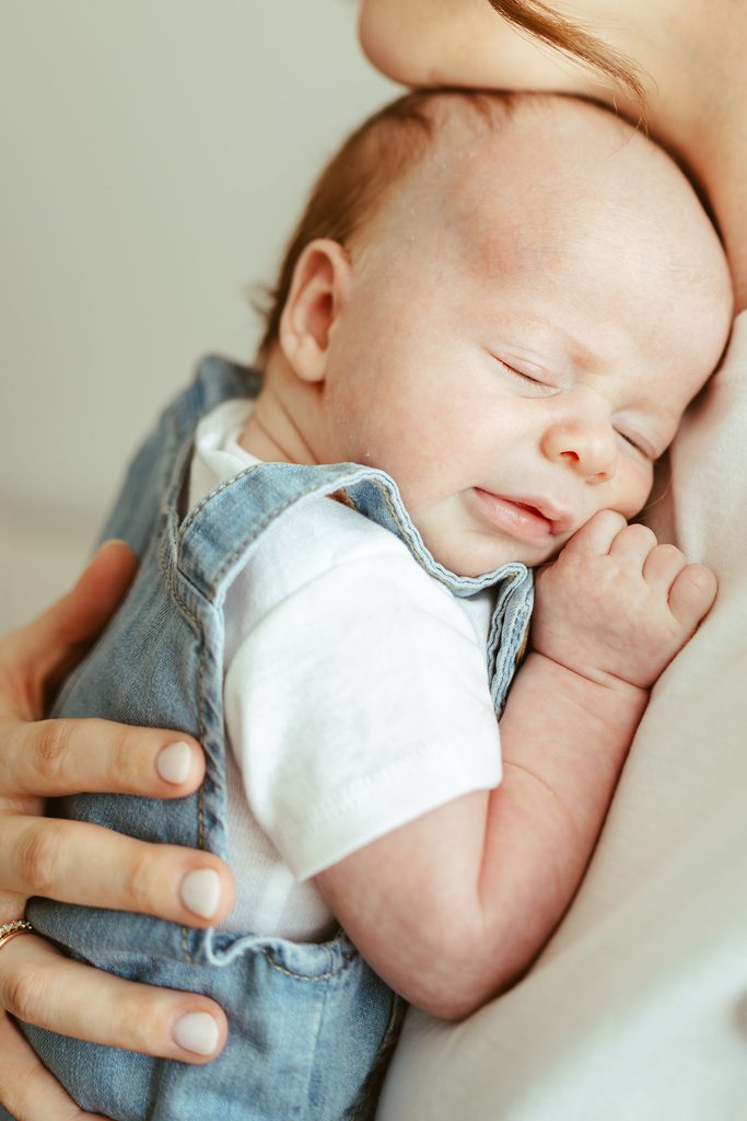 Close-up newborn photography of a sleeping baby in denim overalls being gently held by a parent in Gilbert, Arizona.