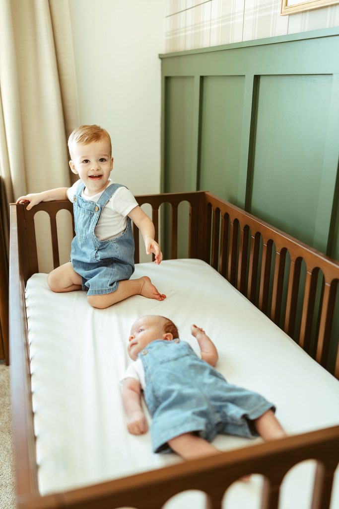 Gilbert sibling newborn photography featuring a toddler sitting beside his newborn sibling in the nursery crib during an in-home session.