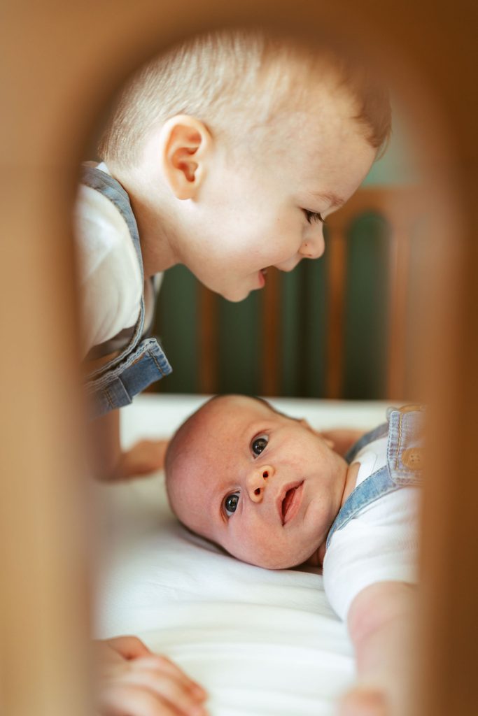 Lifestyle newborn photography in Gilbert, Arizona featuring a toddler leaning over his newborn sibling and looking at the camera in the nursery.