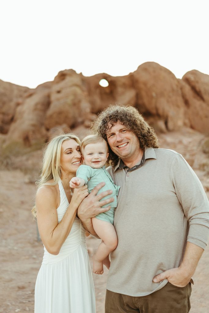 Scottsdale family photographer documenting a lifestyle desert session with parents holding their baby boy next to red rock formations at sunset.