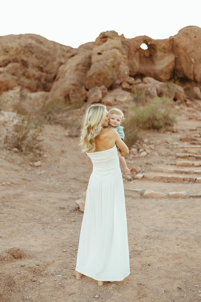 Queen Creek family photography showcasing an editorial, cinematic view of a mother in a white dress carrying her toddler up the paths at Papago Park.