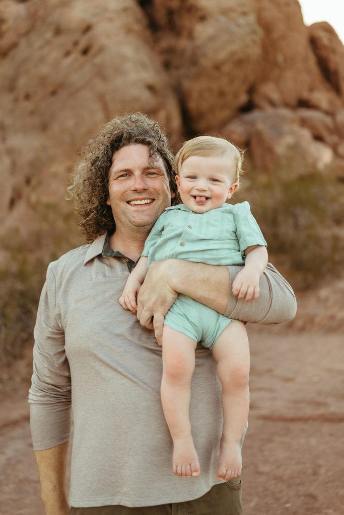 Gilbert family photography featuring a father holding his toddler boy in a sage green outfit against a desert red rock background.