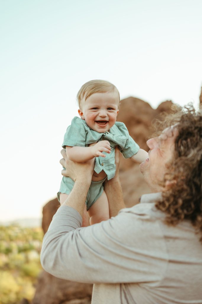 Queen Creek family photography documenting a joyful lifestyle moment as a dad lifts his laughing baby boy up into the golden hour sky.