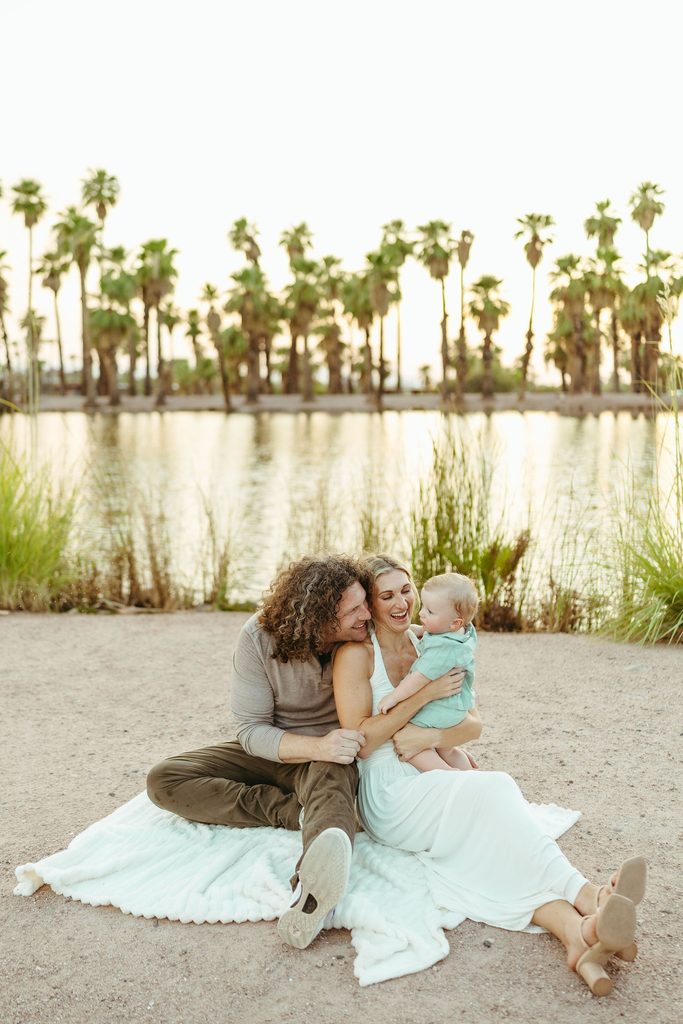 Scottsdale family photographer capturing a relaxed, high-end lifestyle portrait of a family of three snuggling on a white knit blanket by the water.