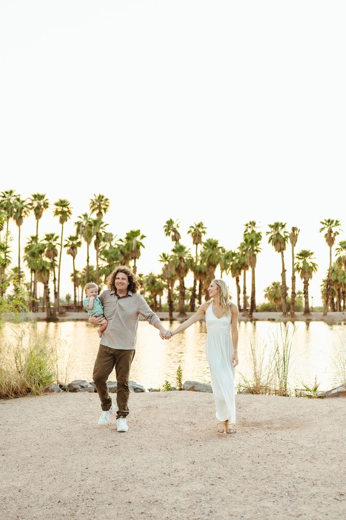 Mesa family photography showcasing an unposed, joyful moment as parents hold hands and walk with their baby boy along a palm tree-lined shore.