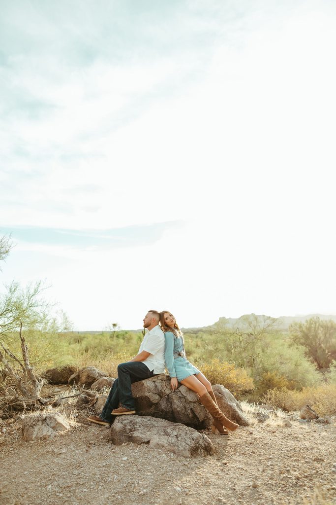 Arizona couples photographer capturing a romantic moment as an engaged couple sits together on rocks surrounded by the Sonoran Desert and Superstition Mountains.