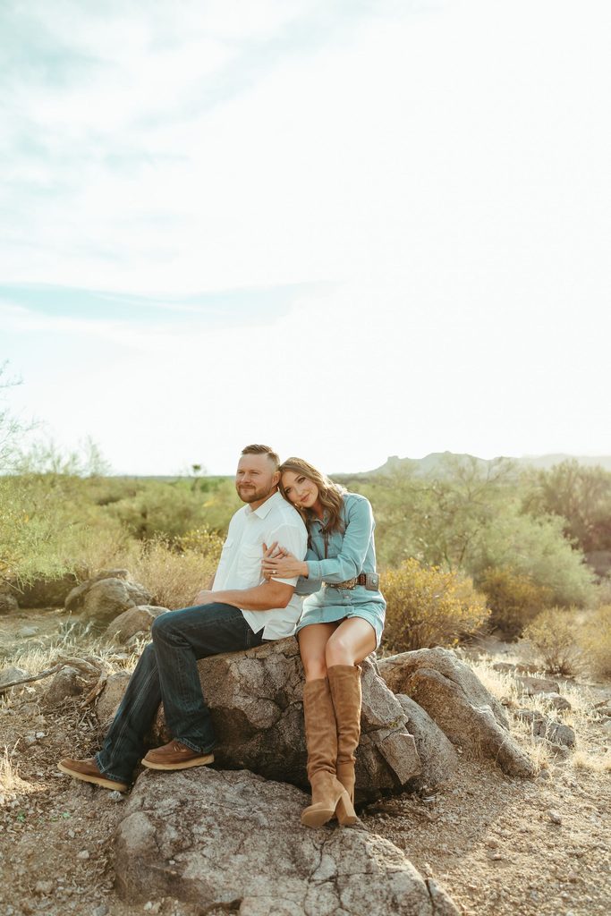 Superstition Mountains engagement photos of a couple sitting closely together on a desert rock surrounded by saguaros and golden Arizona landscape.