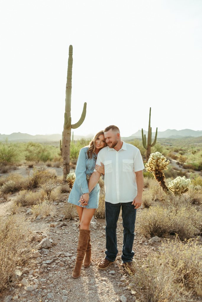 Scottsdale engagement photographer capturing a couple standing together in the Arizona desert with the Superstition Mountains and towering saguaro cacti behind them.
