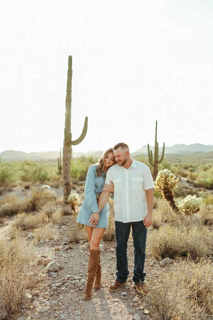 Arizona engagement photographer capturing a couple walking together through the desert beneath towering saguaros with the Superstition Mountains in the background.
