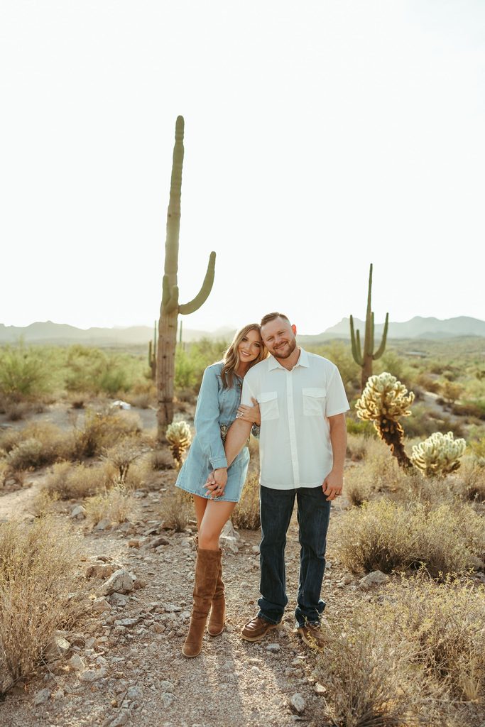 Superstition Mountains engagement session featuring a couple standing together on a desert trail surrounded by golden grasses, saguaros, and mountain views.