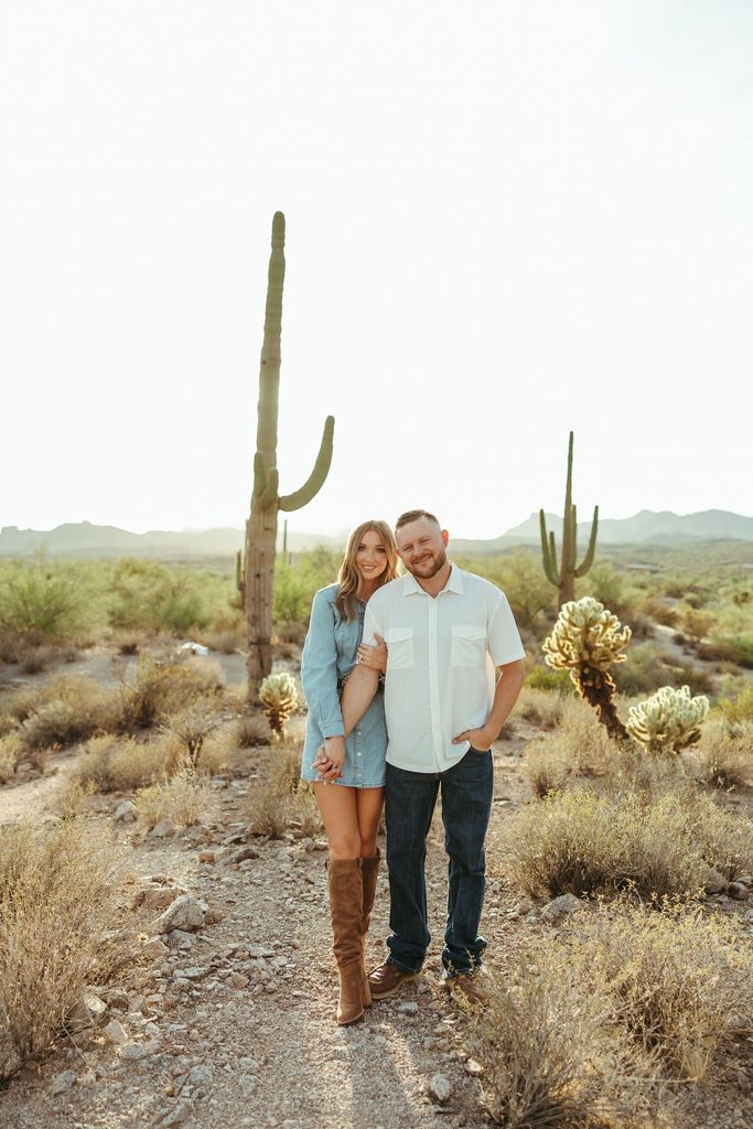 Apache Junction engagement photographer capturing a couple standing together on a desert trail with towering saguaros and the Superstition Mountains behind them.
