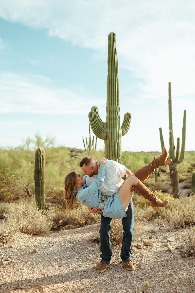Arizona engagement photography featuring a playful couple dipping and laughing together on a desert trail surrounded by saguaros near the Superstition Mountains.